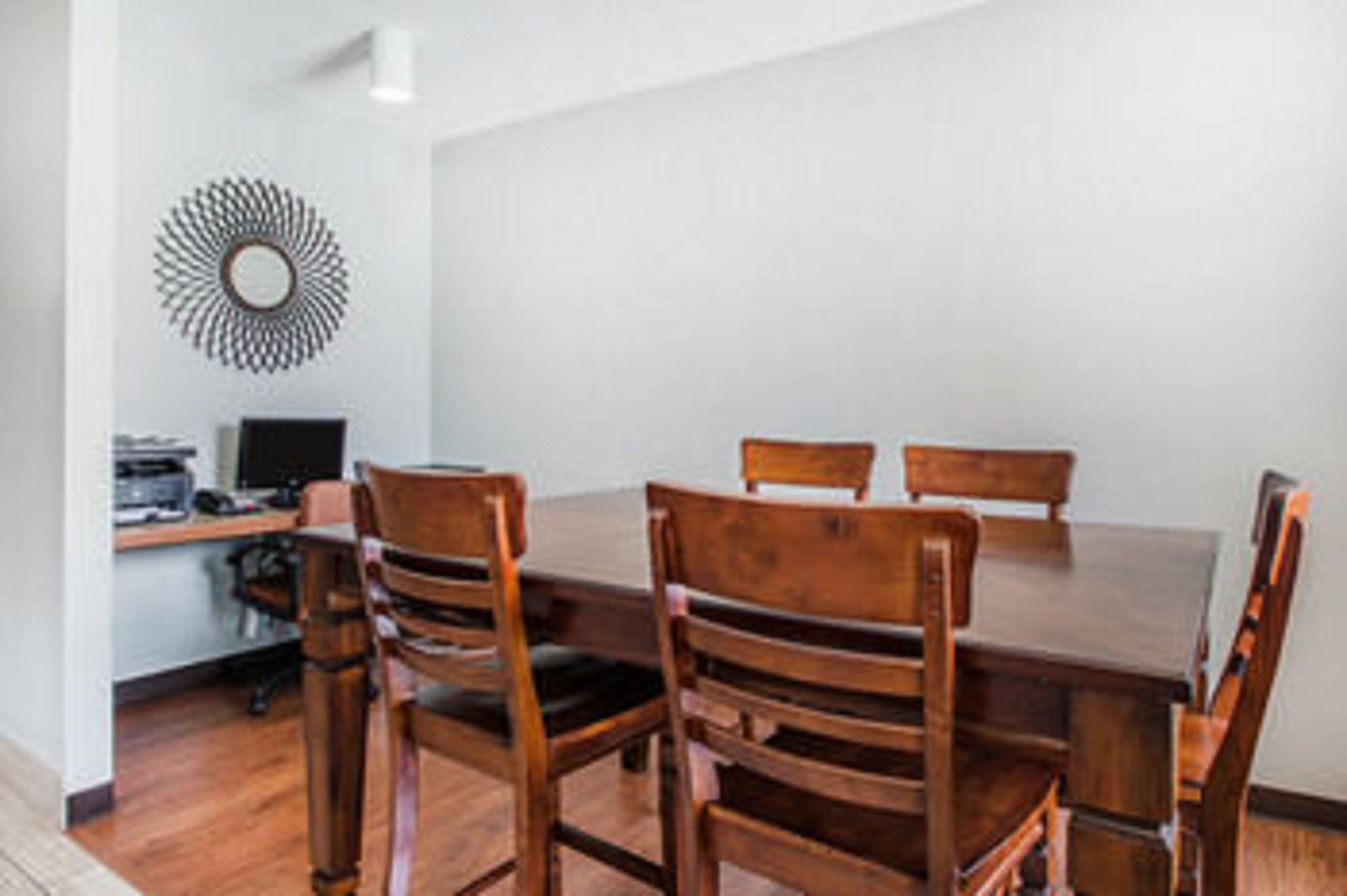 Un comedor con una mesa y sillas de madera, un espejo decorativo colgado en la pared y un escritorio pequeño con un ordenador e impresora.