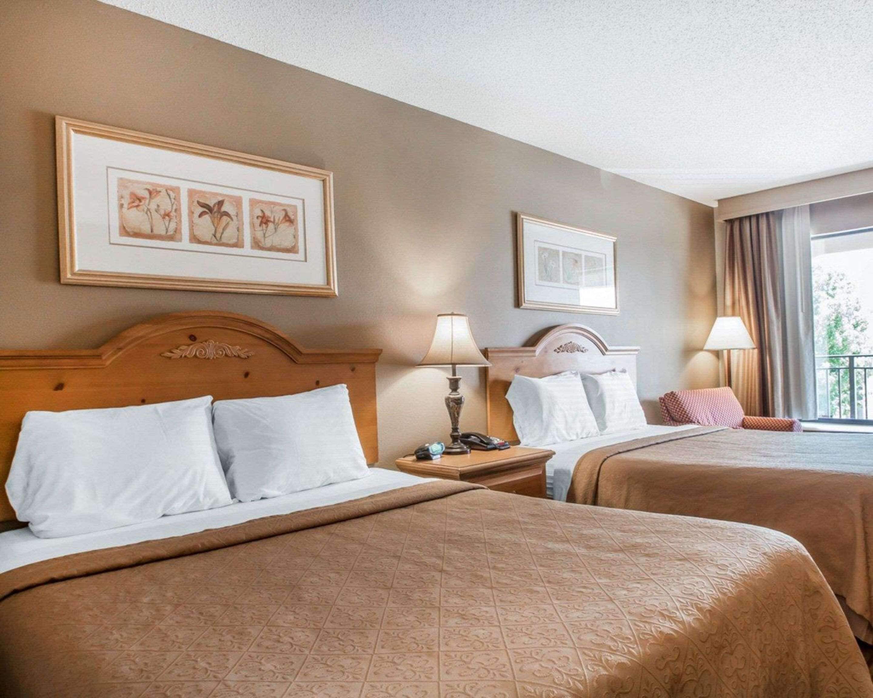 Een hotelkamer met twee bedden, elk met een houten hoofdeinde, witte lakens en een bruin geborduurd sprei. Er hangen foto's in lijsten aan de muur, een nachtkastje met een lamp en een raam met gordijnen.