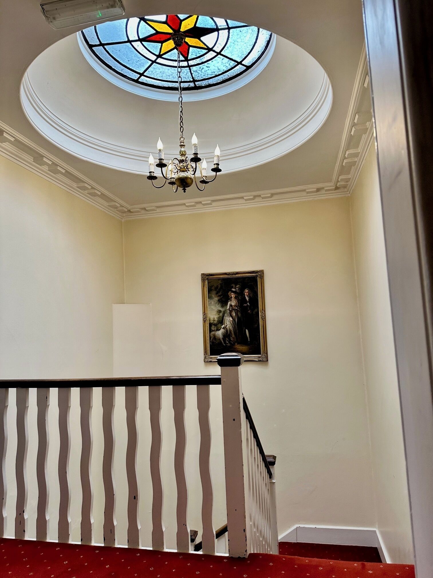 Uma escadaria com corrimão branco e tapete vermelho. Um lustre pendurado no teto e um quadro na parede.