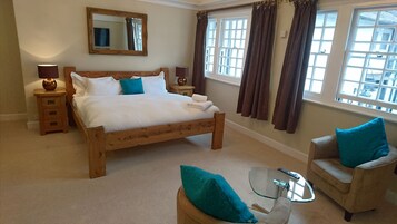 Quarto com cama de madeira, duas mesas de cabeceira, uma mesa de centro de vidro, uma cadeira e uma janela grande com cortinas.