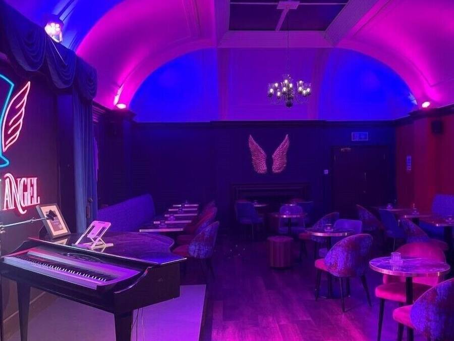 A room with a piano, tables, chairs, and a neon sign.