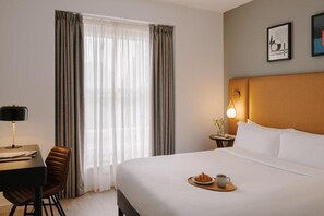 Une chambre d’hôtel avec un lit, un bureau et une chaise, une lampe et une fenêtre avec des rideaux.