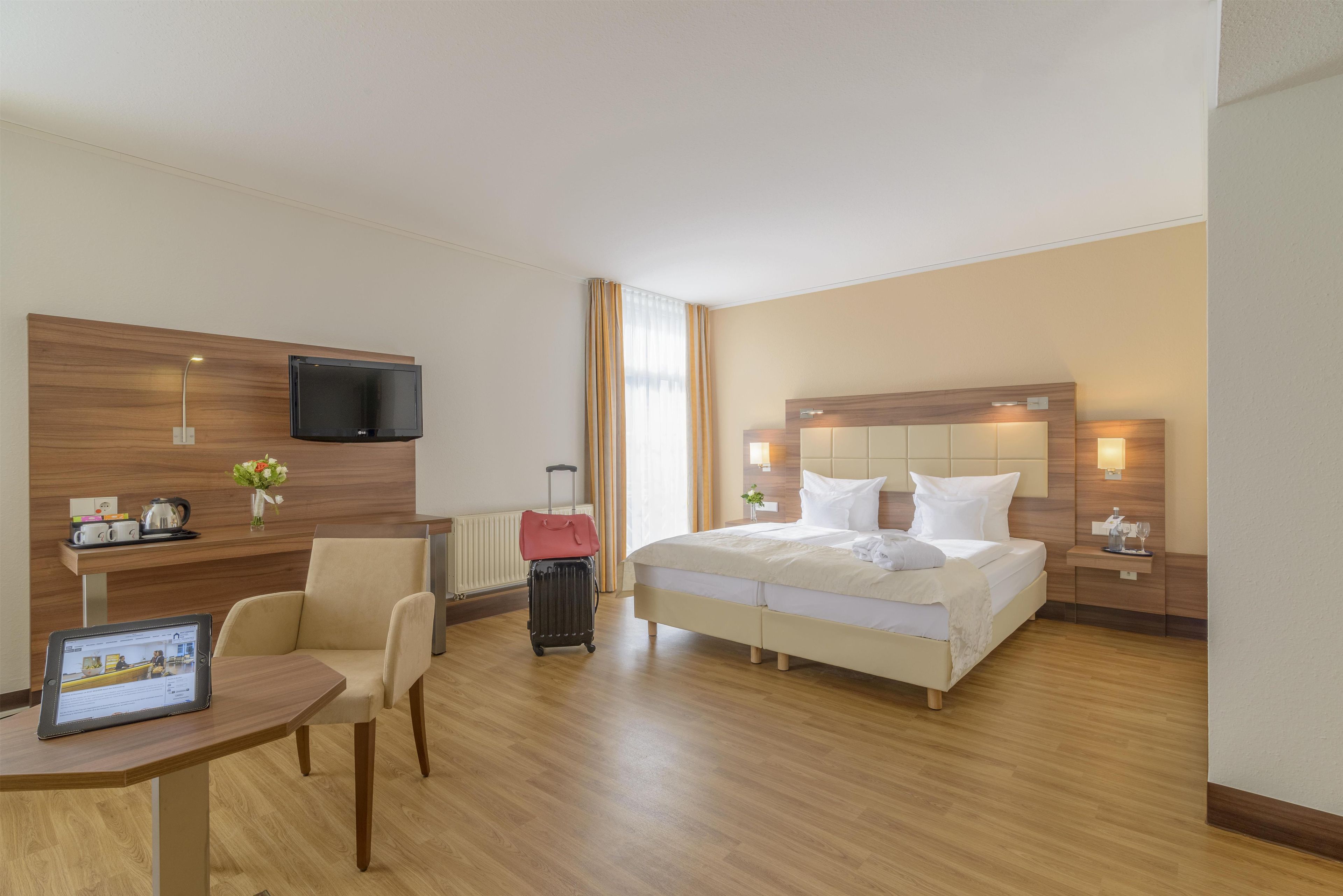 Una habitación de hotel con cama, mesitas de noche, un escritorio con silla, televisor y ventana con cortinas.