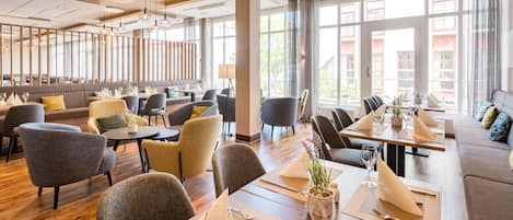 Um restaurante espaçoso com pé-direito alto, amplas janelas e diversas opções de assentos, incluindo mesas e sofás.