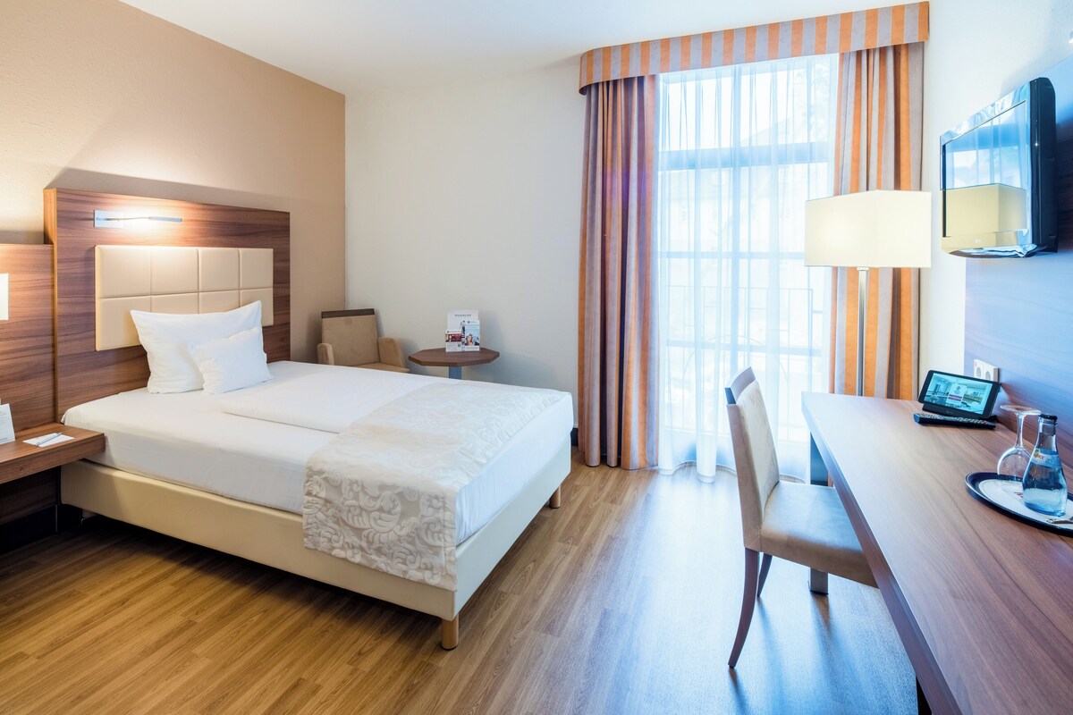 A hotel room with a bed, bedside table, lamp, desk, chair, and a TV.