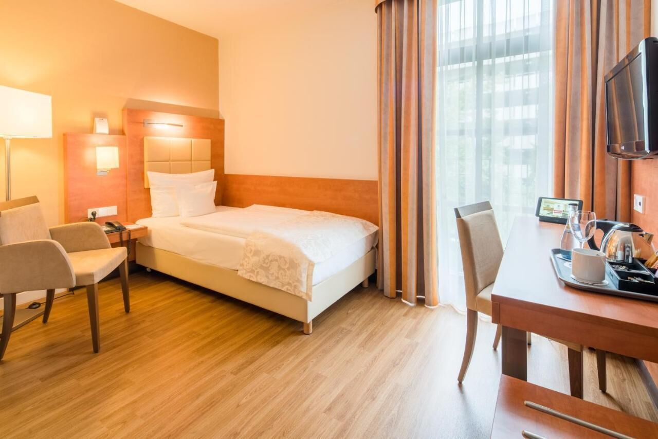 A hotel room with a bed, a desk with a chair, a television, and a window with curtains.
