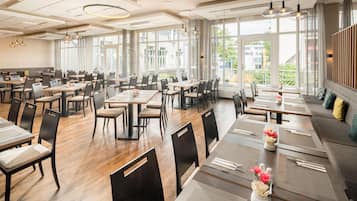 A spacious restaurant with multiple dining tables and chairs, large windows offering views of the outside, and modern pendant lighting.