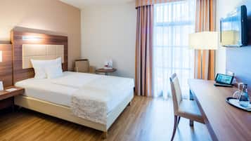 Quarto de hotel com cama, mesa de cabeceira, abajur, cadeira, escrivaninha e televisão.
