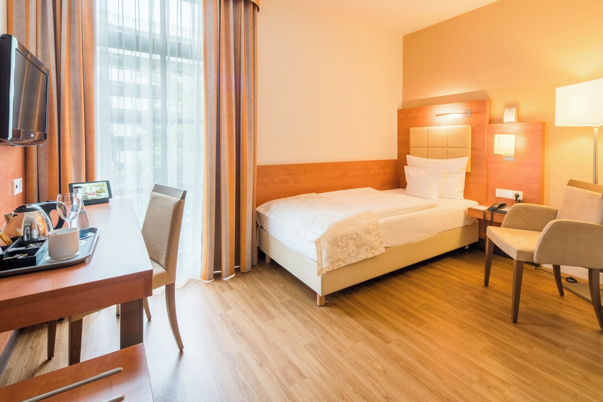 A hotel room with a bed, bedside table, chair, desk, and a television.