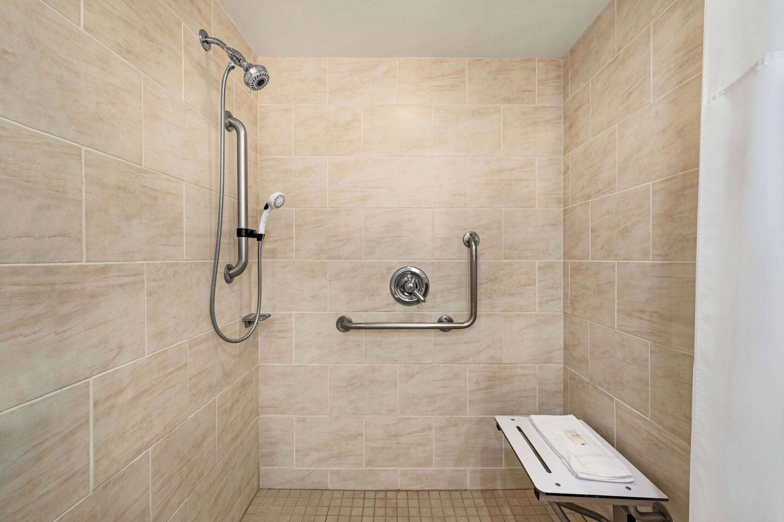 A shower area with beige tiles, a handheld showerhead, a wall-mounted faucet, and a grab bar.