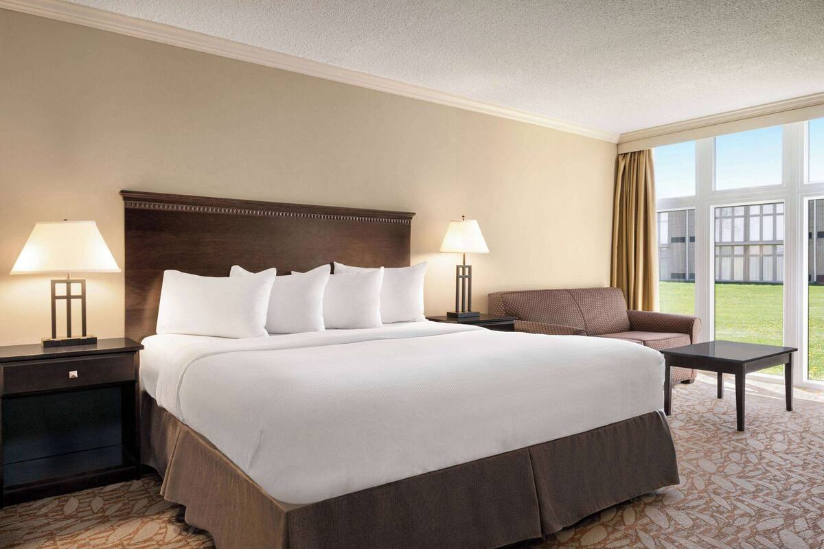 Habitación de hotel con una cama grande, dos mesitas de noche con lámparas, un sofá marrón y vistas al exterior a través de amplias ventanas.