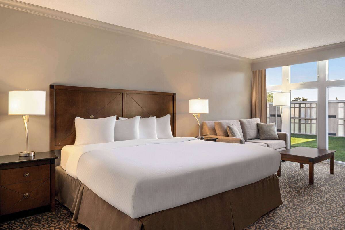 Habitación de hotel con una cama grande, dos mesitas de noche con lámparas, un sofá y vistas a un espacio verde exterior.