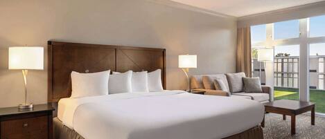 A hotel room with a large bed, two bedside tables with lamps, a sofa, and a view of a green outdoor area.