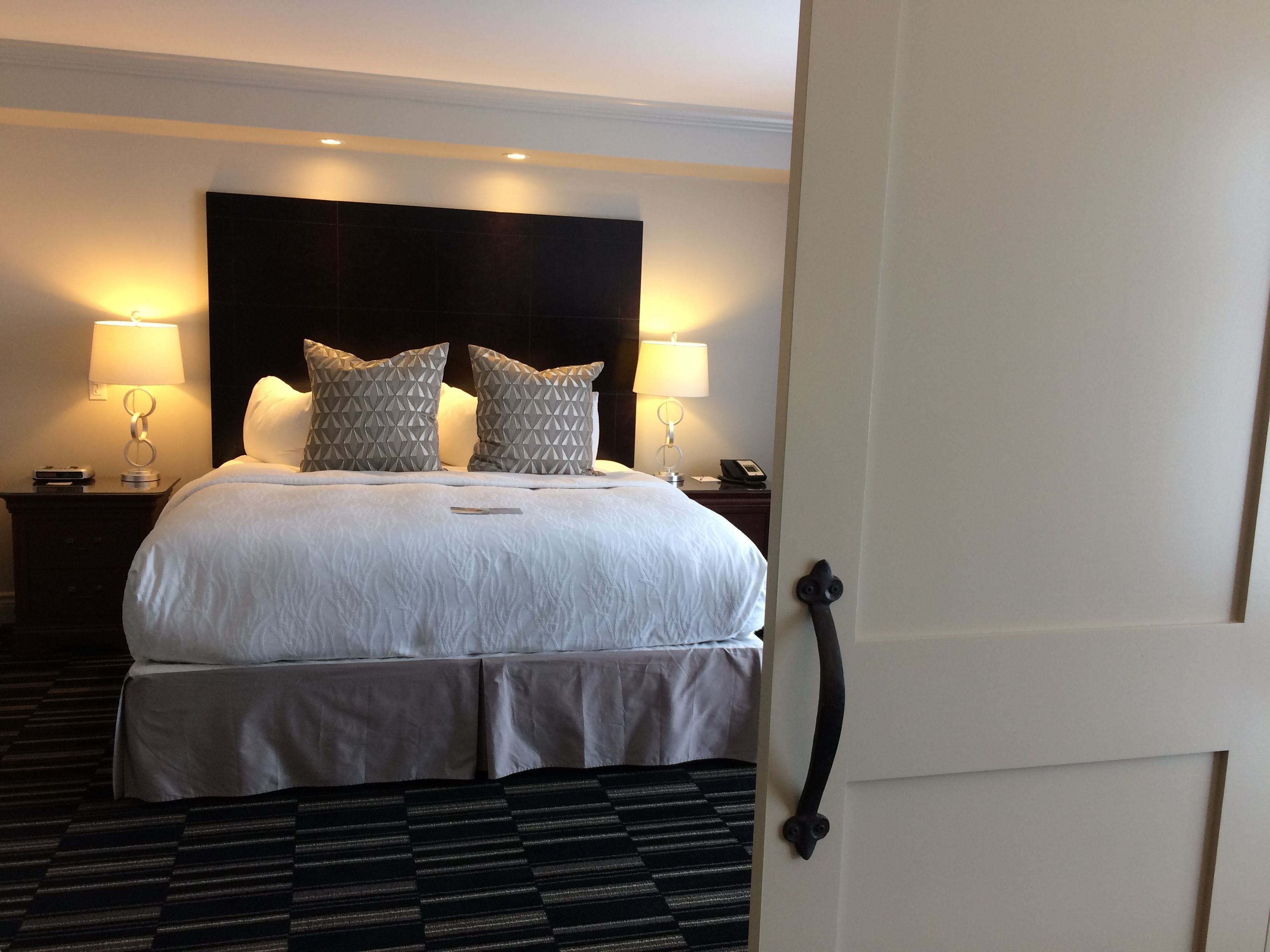 A hotel room with a large bed, two bedside lamps, a headboard, a bedside table with a phone, and a patterned carpet.