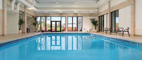 An indoor swimming pool with clear glass walls, a tiled floor, and ceiling lights.