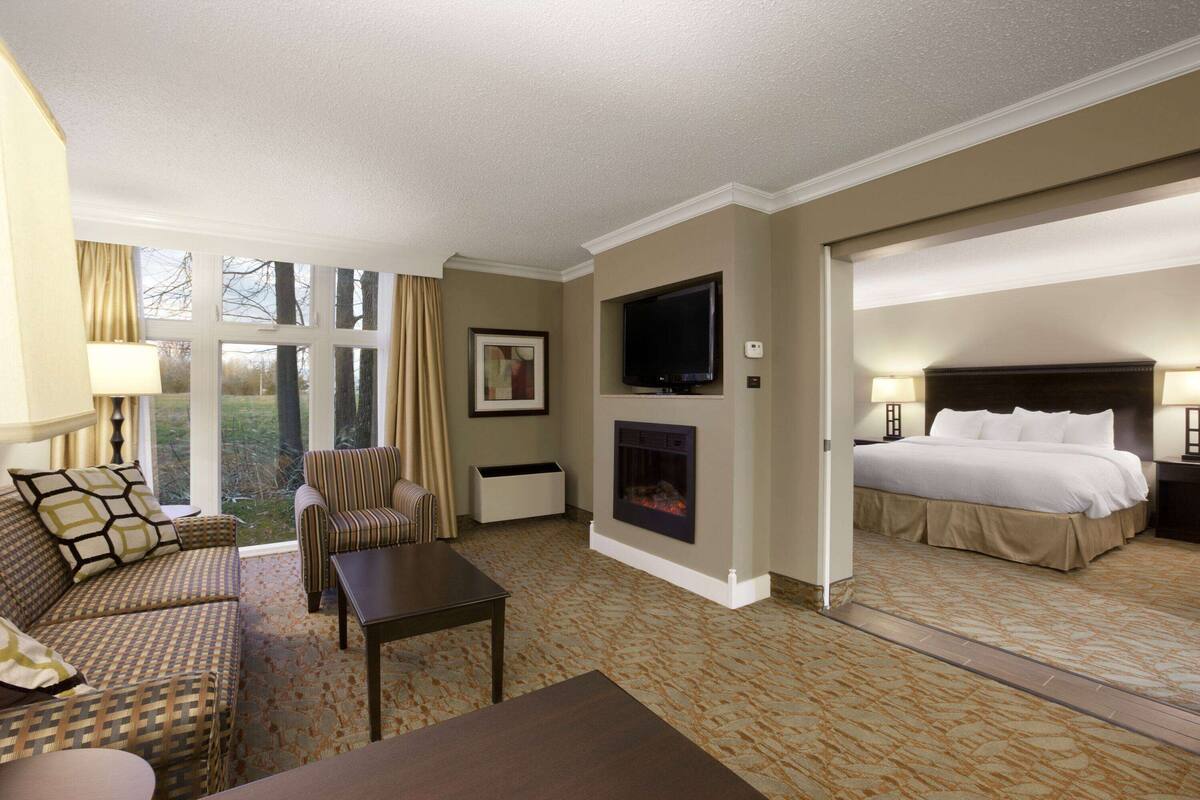 Habitación de hotel con sofá, sillón, televisor de pantalla plana montado en la pared, chimenea, cama y mesita de noche con lámpara.