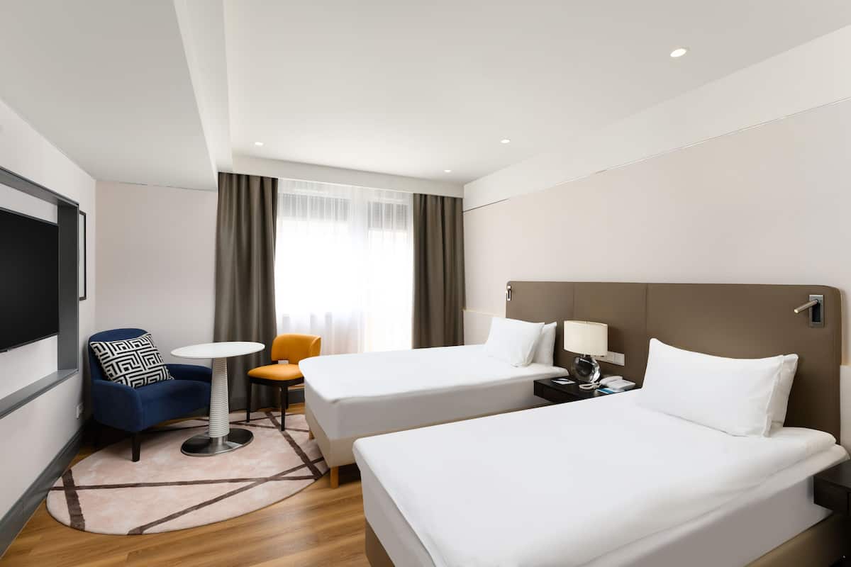 Een hotelkamer met twee eenpersoonsbedden, een klein rond tafeltje, een blauwe fauteuil, een stoel en een televisie.