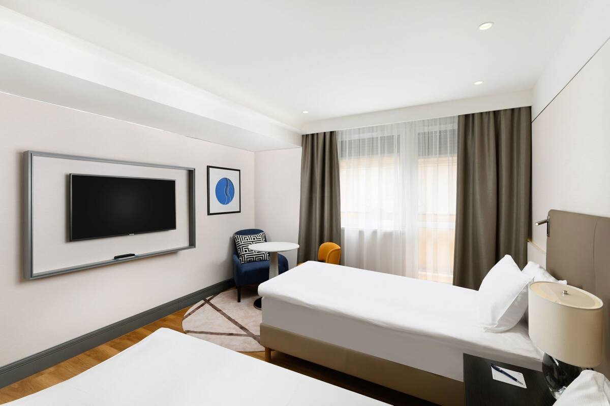 Une chambre d’hôtel avec un lit, un bureau, une chaise, une télévision et une fenêtre avec des rideaux.