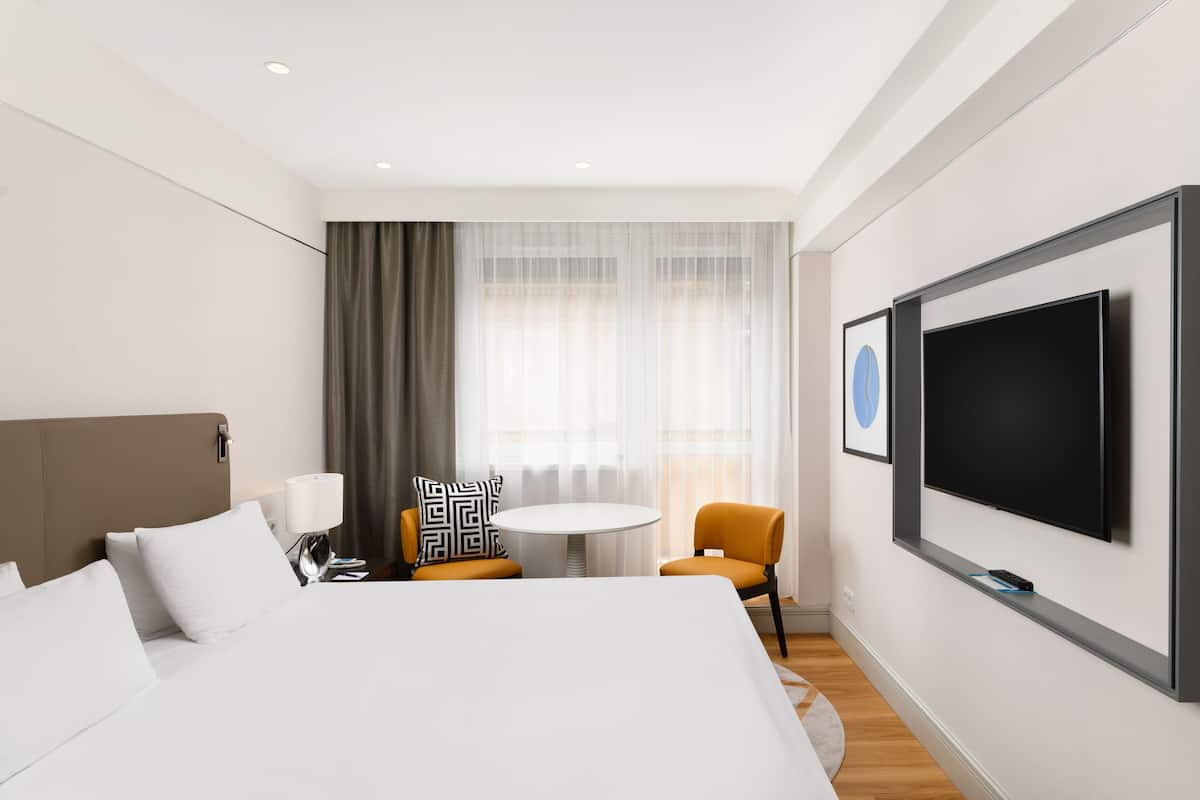 Een hotelkamer met een groot bed, een aan de muur bevestigde televisie, een ronde tafel, twee stoelen en een raam met gordijnen.
