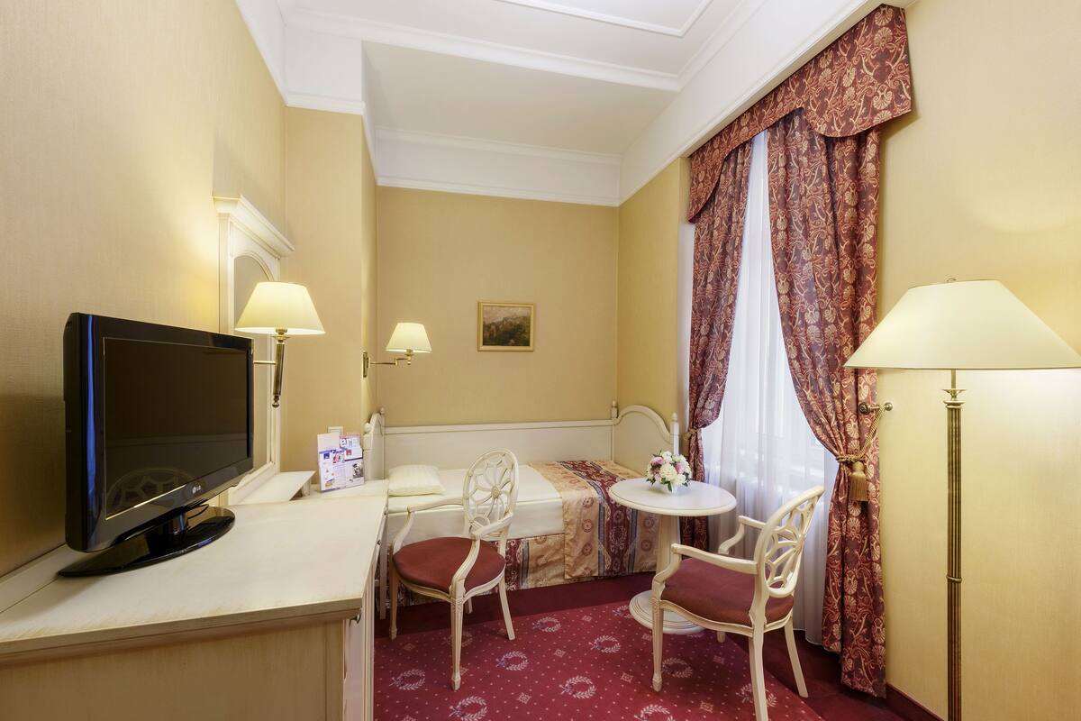 Een hotelkamer met een bed, een bureau met een computer monitor, twee stoelen, een tafeltje met een bloevas en twee lampen.