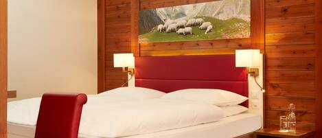 A hotel room with a wooden paneled wall, a red upholstered headboard, a white bed with white linens, two bedside lamps, and a painting of sheep on a mountain.