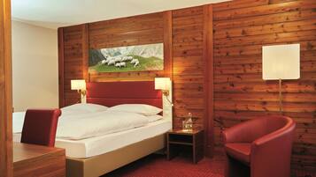 Ein Hotelzimmer mit einer holzverkleideten Wand, einem Bett mit weißer Bettwäsche, einem rot gepolsterten Sessel, einem kleinen Nachttisch mit Lampe und einem Bild von Schafen in den Bergen.