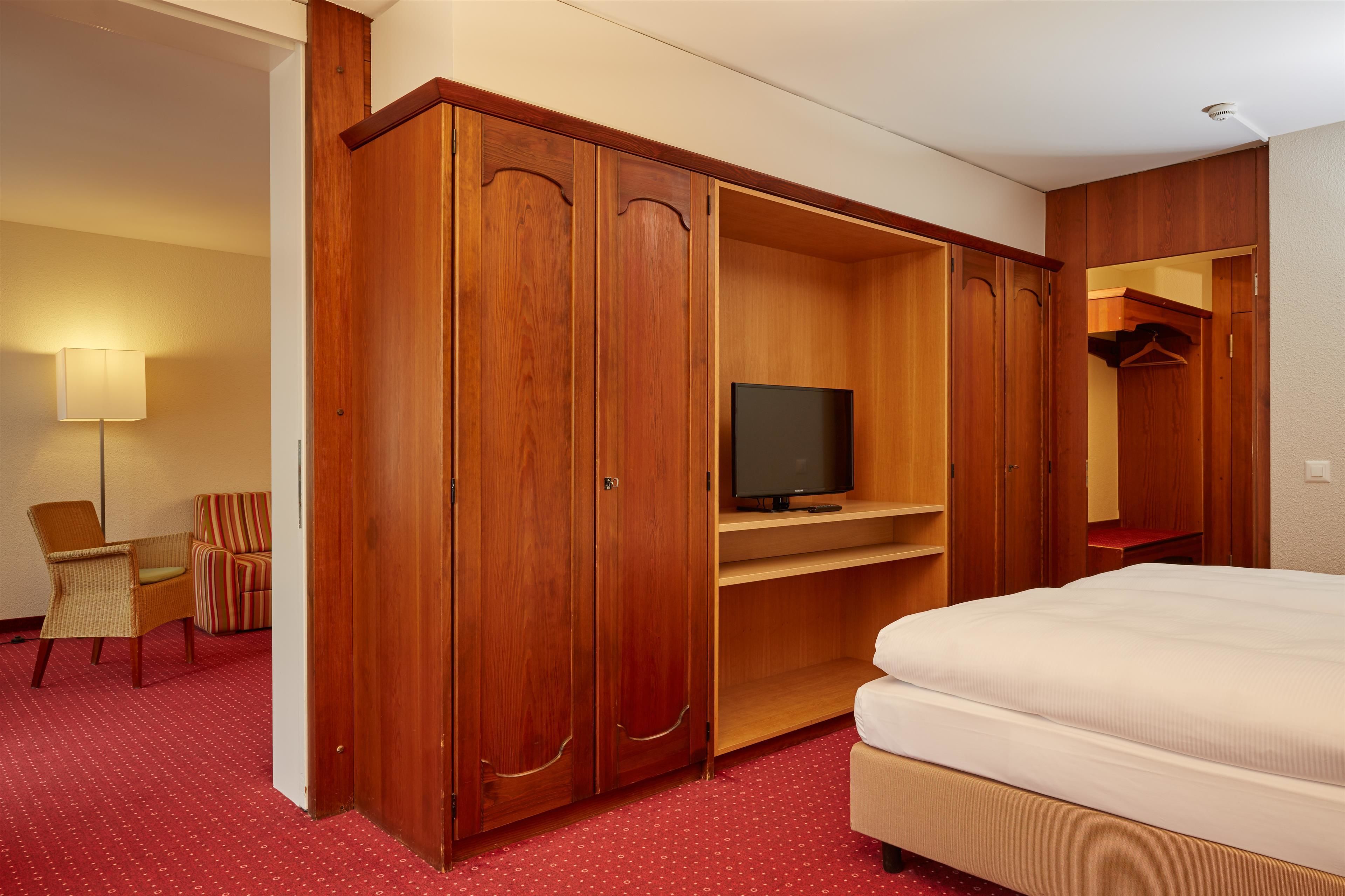 A hotel room with a large wooden wardrobe, a television on a shelf, a bed, a chair, and a lamp.