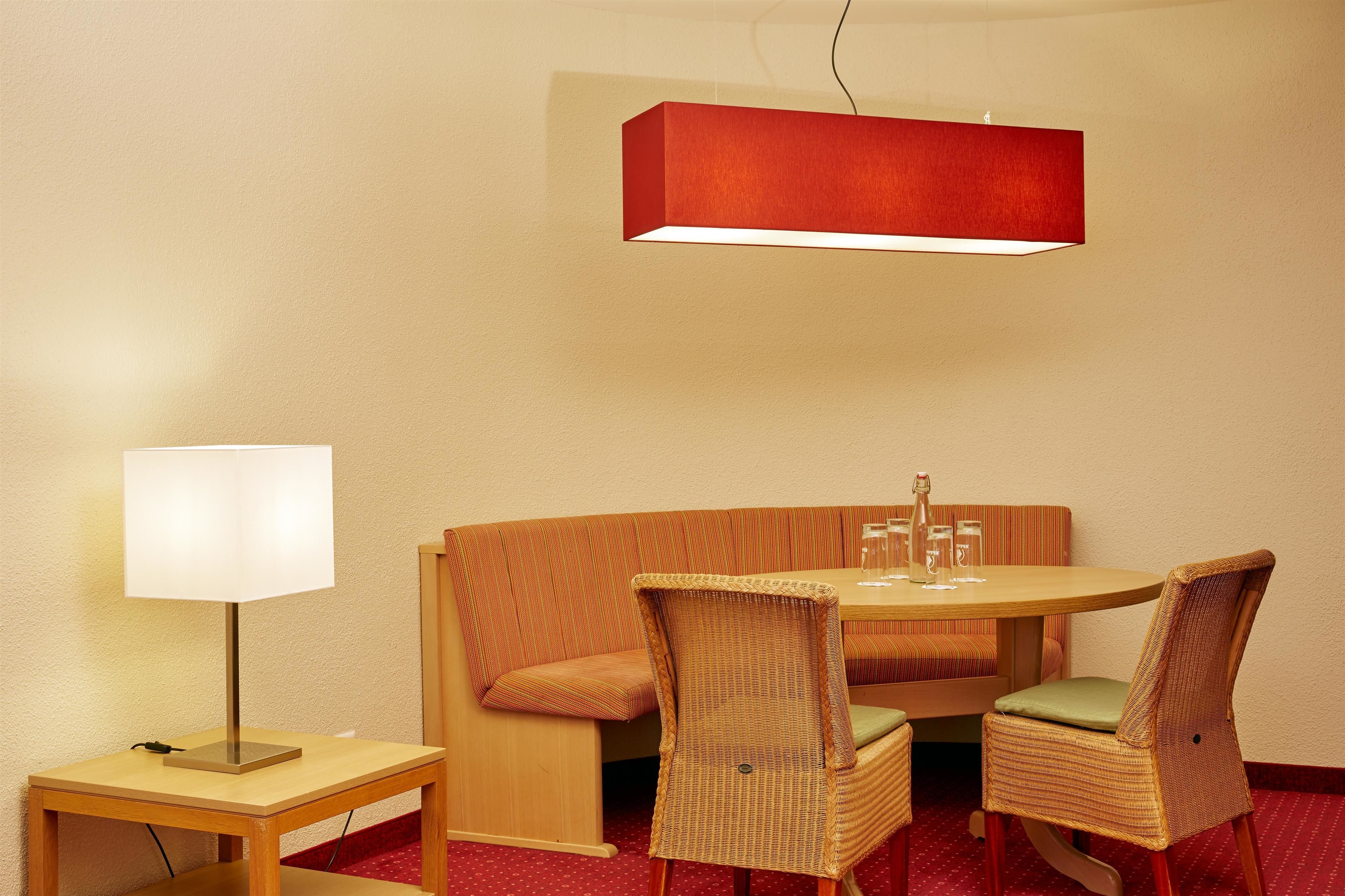 Una habitación de esquina con una mesa de madera, dos sillas de mimbre, un banco y una lámpara roja fijada a la pared.