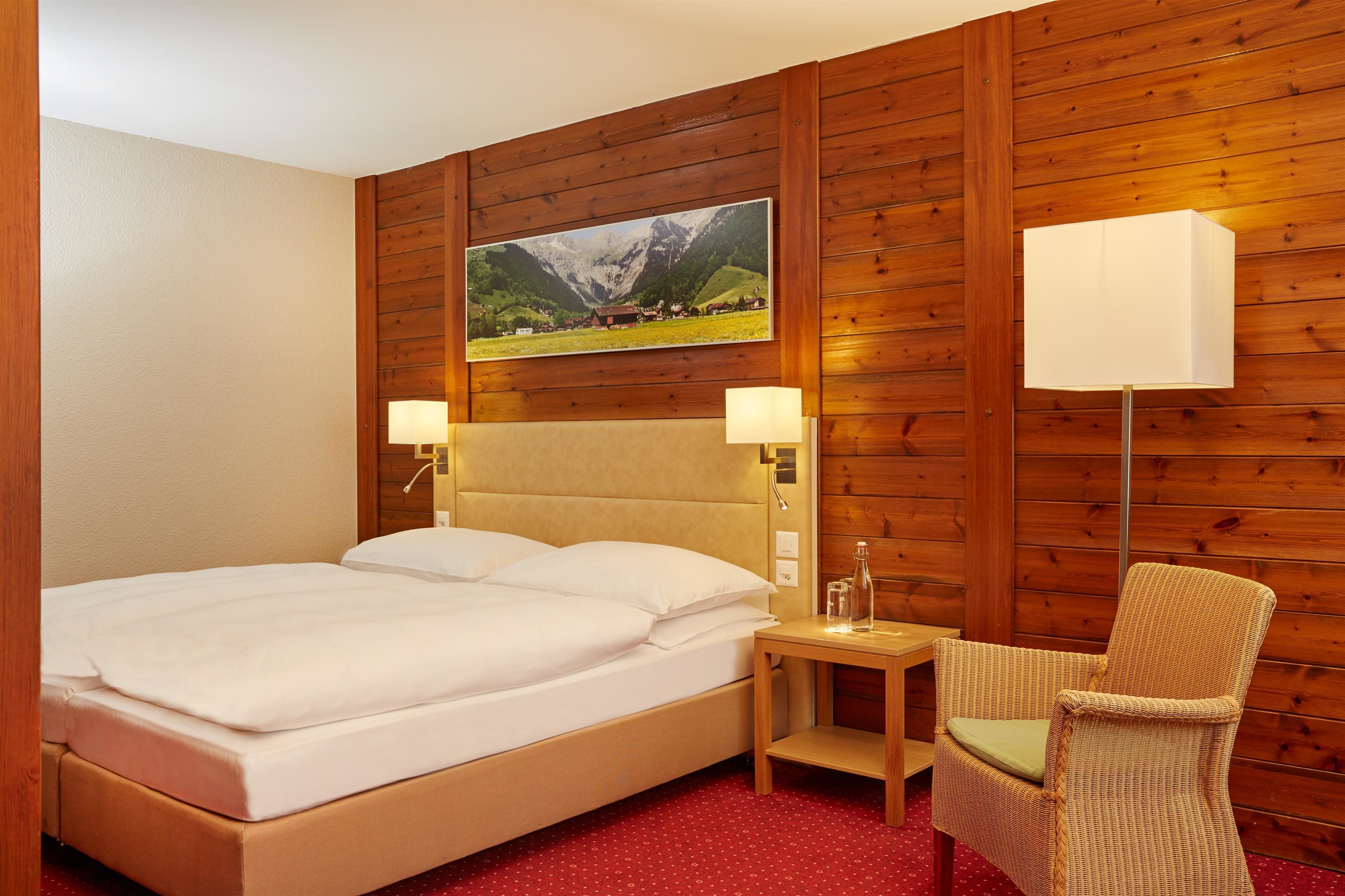 Una habitación de hotel con pared de paneles de madera, una cama grande con ropa blanca, dos lámparas de noche, una mesita de noche de madera y una silla de mimbre.