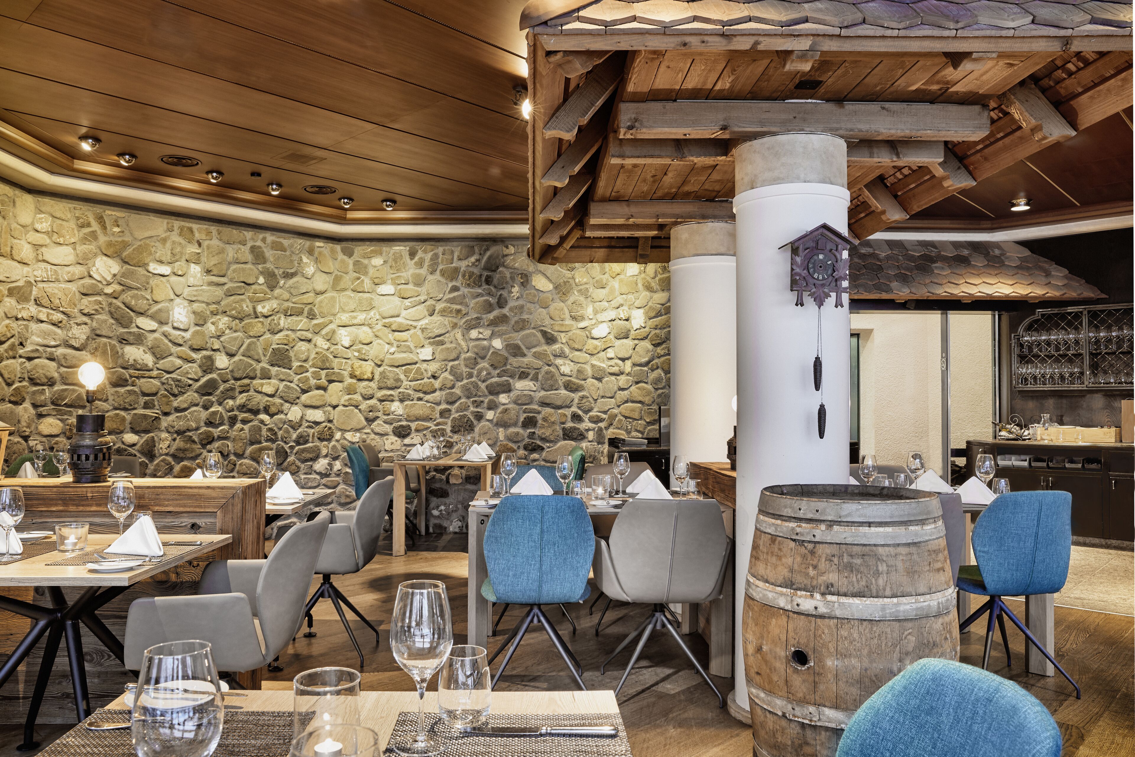 A restaurant with stone walls, wooden ceiling beams, and rustic decor including a barrel. Tables are set with wine glasses and napkins.