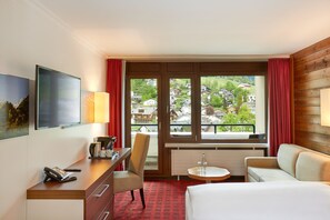 A hotel room with a desk, chair, bed, and a view of a town through large windows.