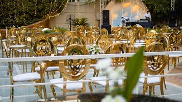 Uma sala de banquetes com mesas redondas e cadeiras, um palco com um cenário e luzes de corda.