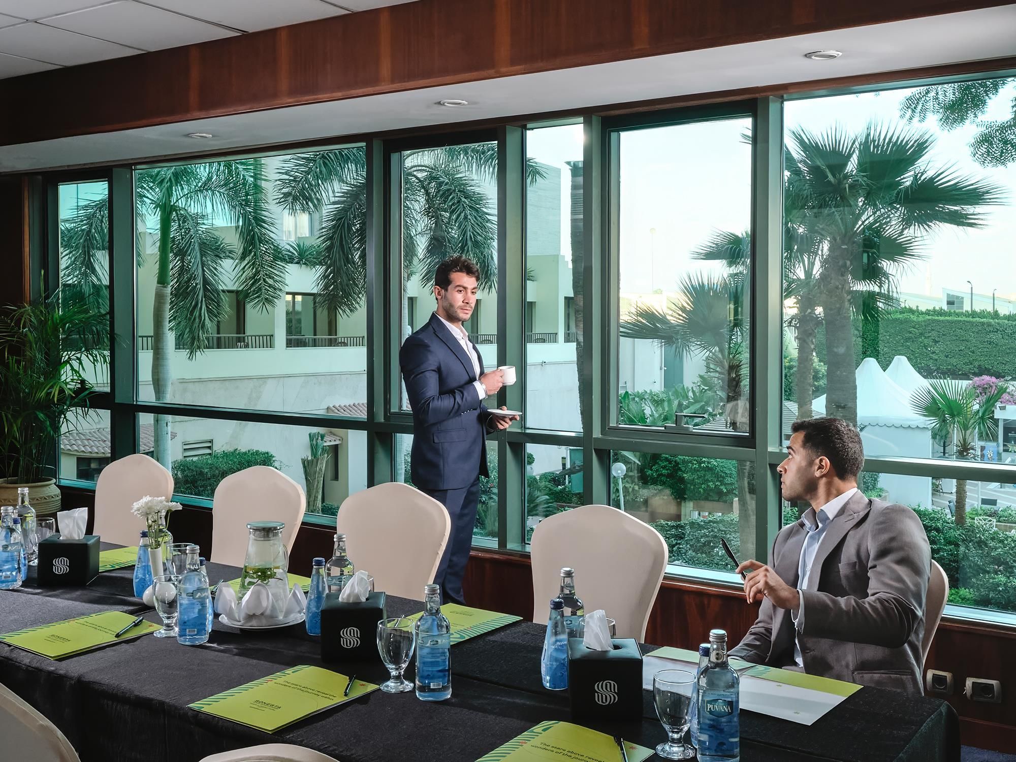 A conference room with a long table set for a meeting, large windows with a view of palm trees, and a modern interior design.