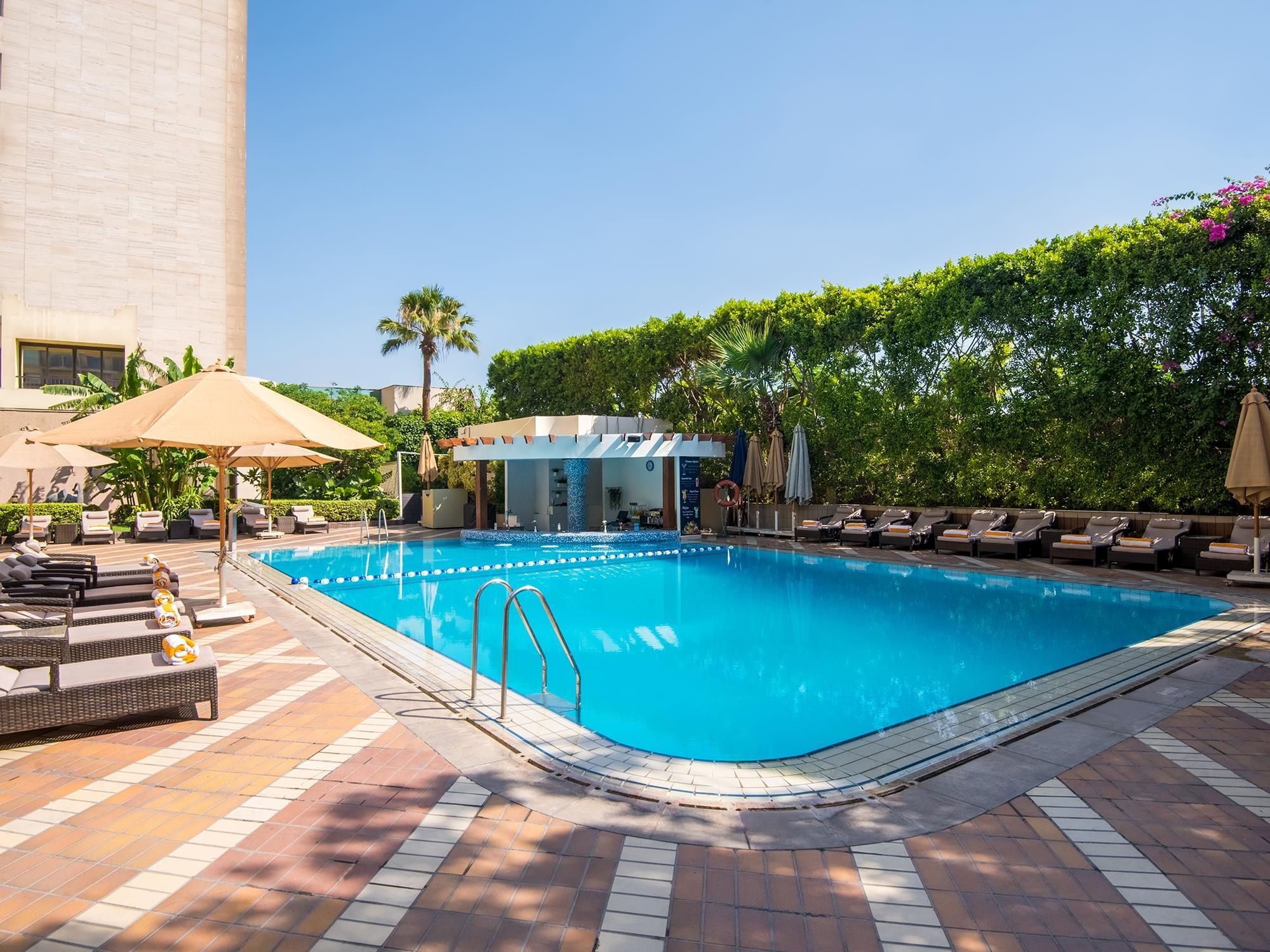 A swimming pool with lounge chairs and umbrellas, surrounded by greenery and a building.