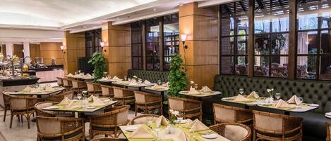 A hotel dining area with multiple tables set for guests, featuring wicker chairs and a green upholstered bench.