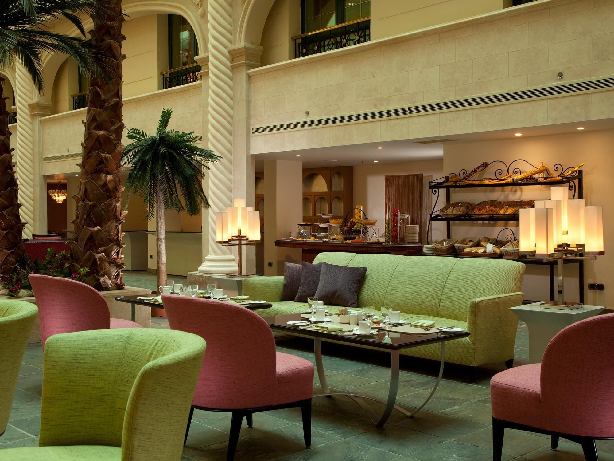 A spacious lobby with a green sofa, multiple chairs, a dining table set for two, and a buffet area with various food items.