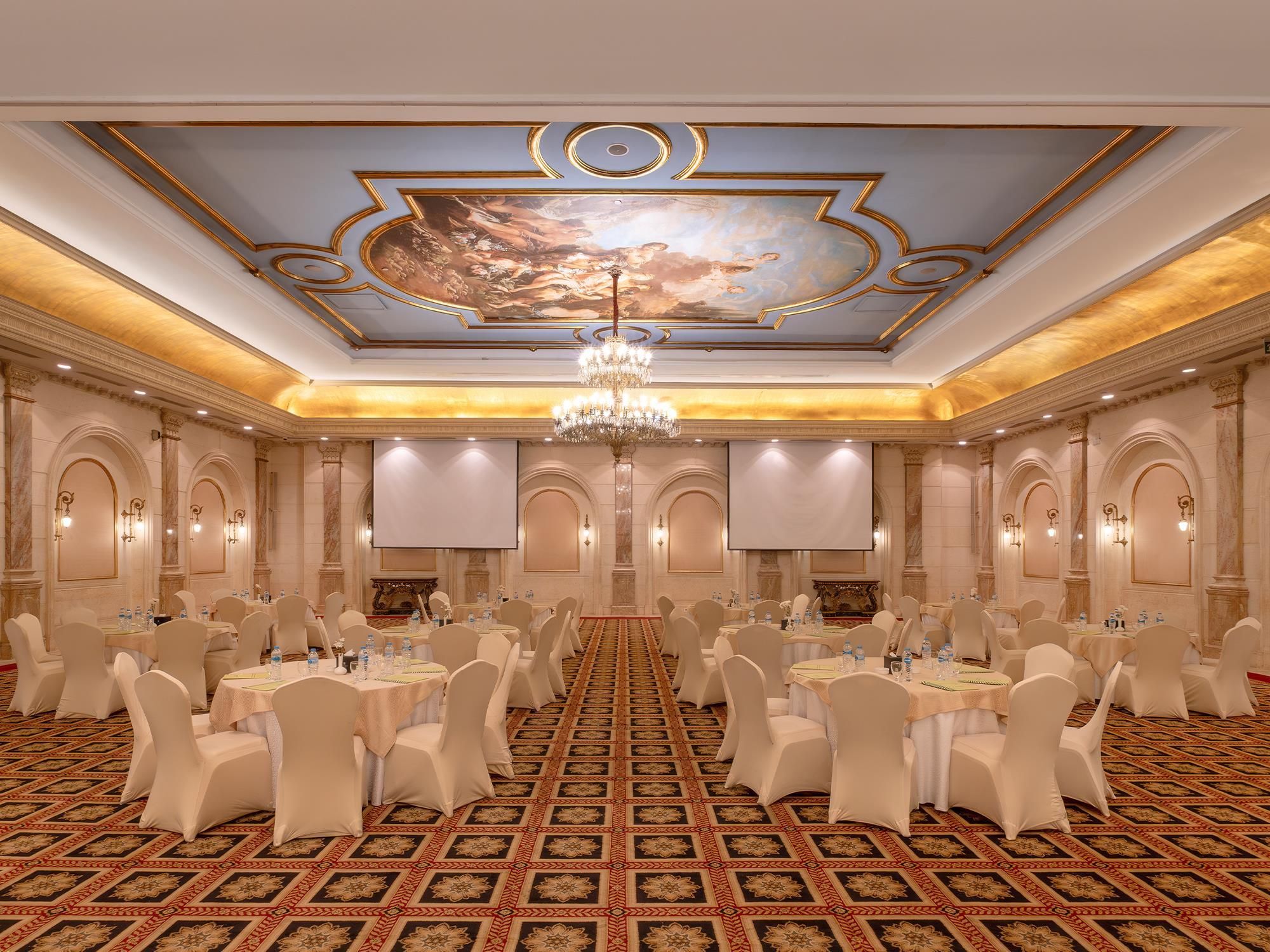 A banquet hall with round tables, white chairs, a chandelier, and a large mural on the ceiling.