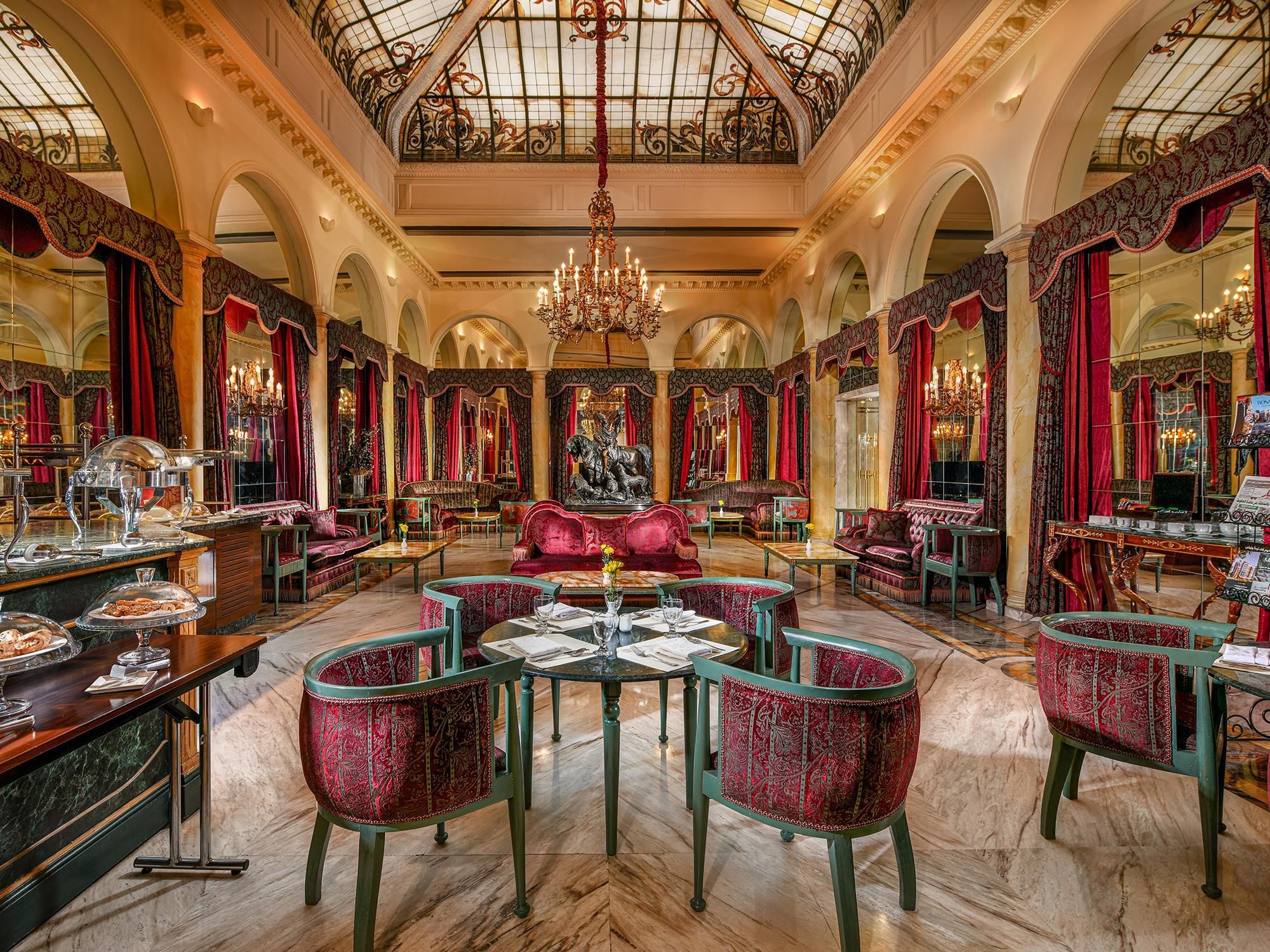 A grand interior with ornate chandeliers, red velvet furniture, and a central statue.
