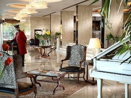 A grand lobby with a piano, patterned chairs, a reception desk, and a large chandelier.