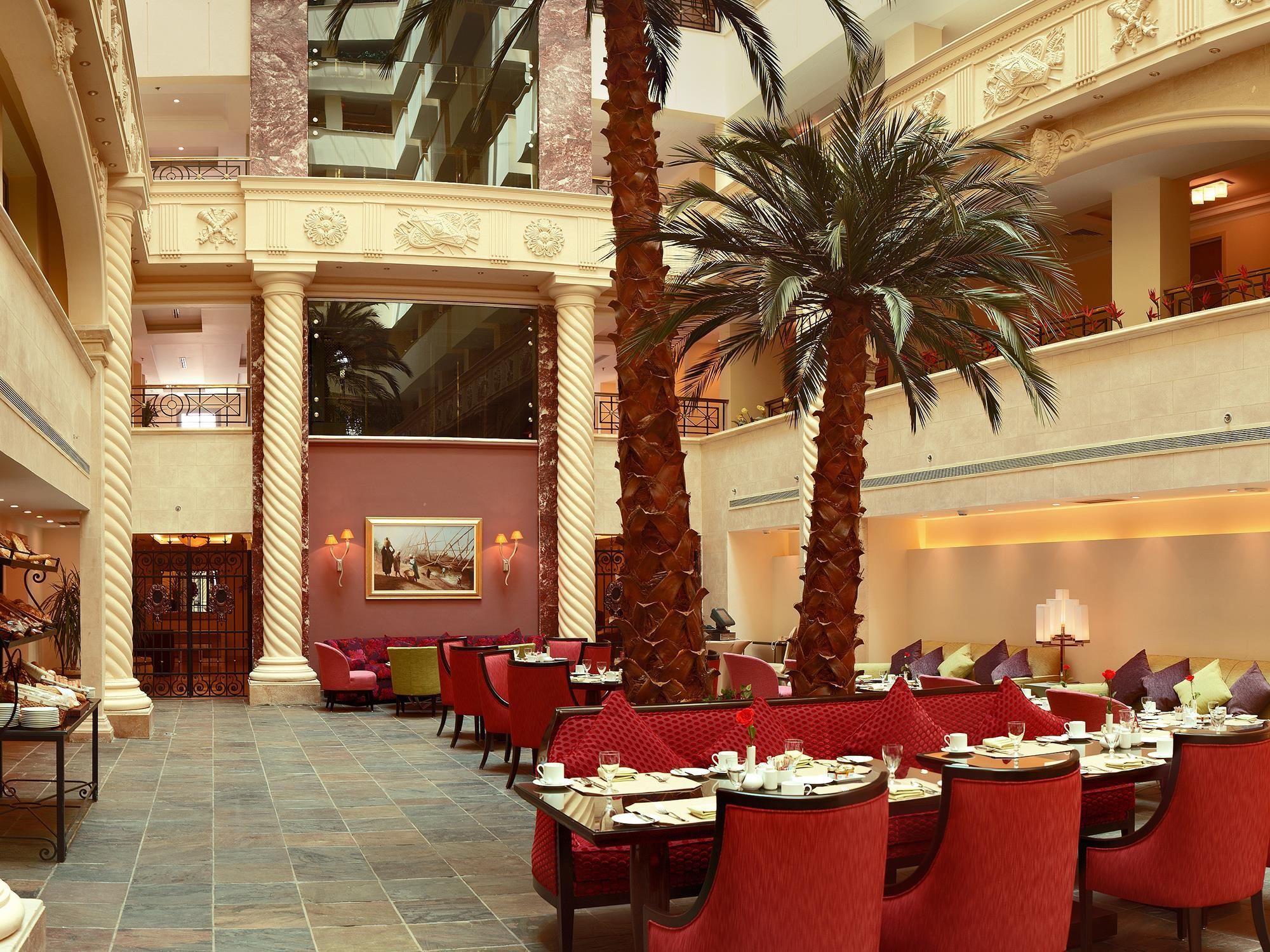 A spacious hotel lobby with high ceilings, a central palm tree, and a dining area with red upholstered chairs and tables.