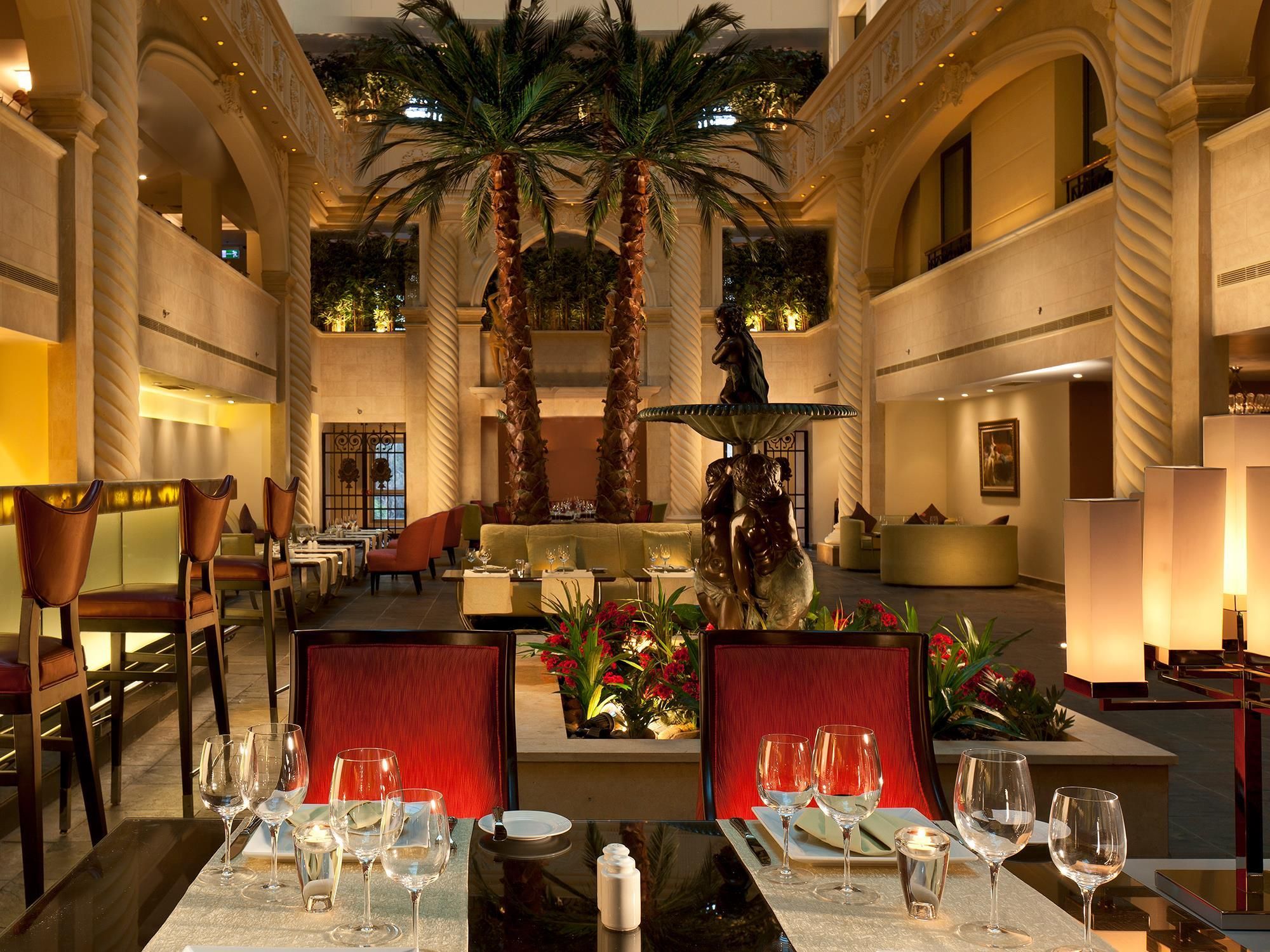 A hotel lobby with a central fountain, palm trees, and a dining area with tables set for guests.