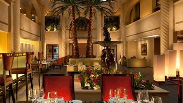 Un vestíbulo de hotel con una fuente central, palmeras y un área de comedor con mesas dispuestas para los huéspedes.