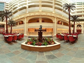 A hotel lobby with a central fountain, multiple dining areas with red chairs, and a high ceiling with balconies.