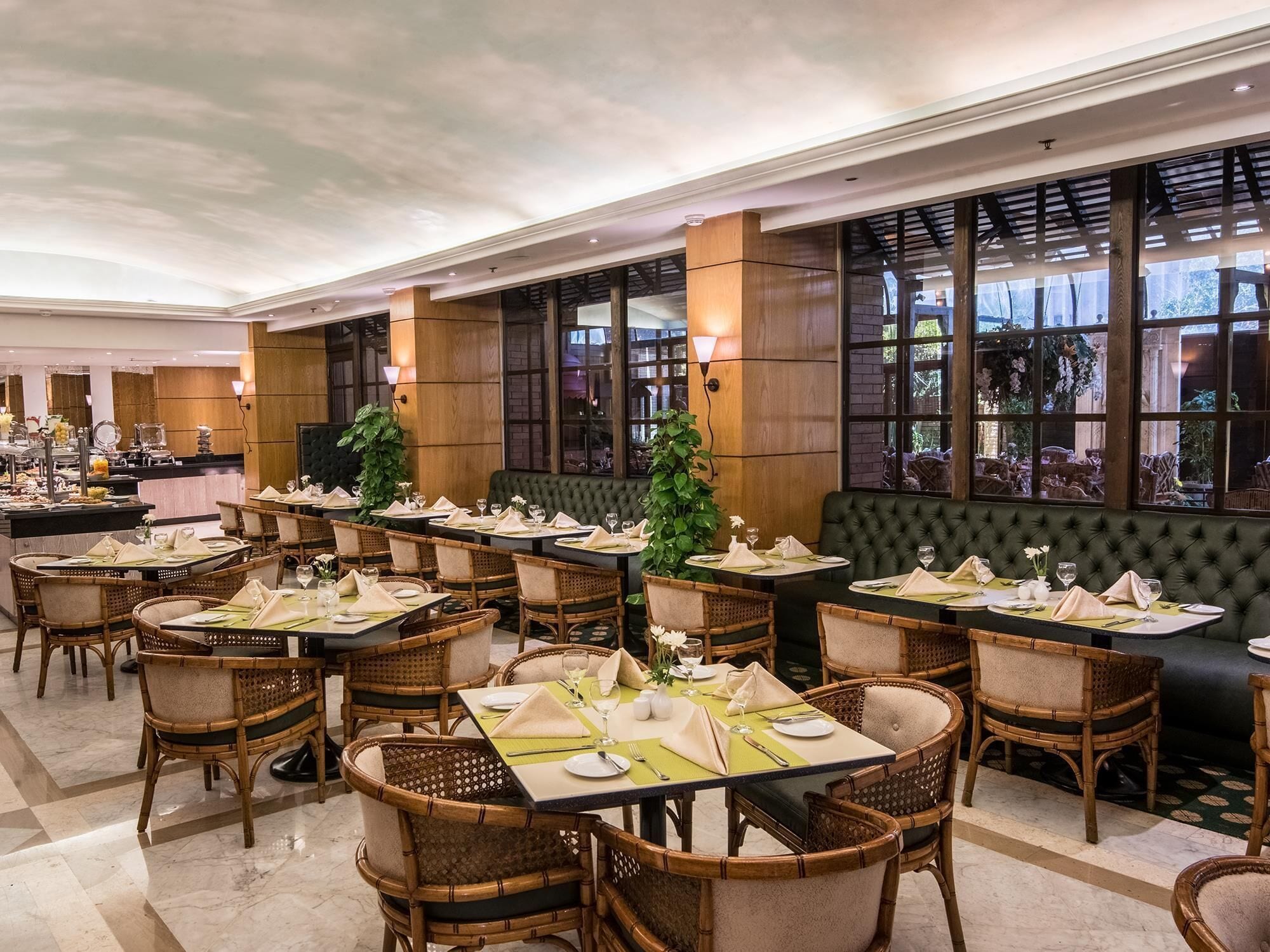 Un comedor de hotel con varias mesas dispuestas para los huéspedes, con sillas de mimbre y un largo banco tapizado de verde.