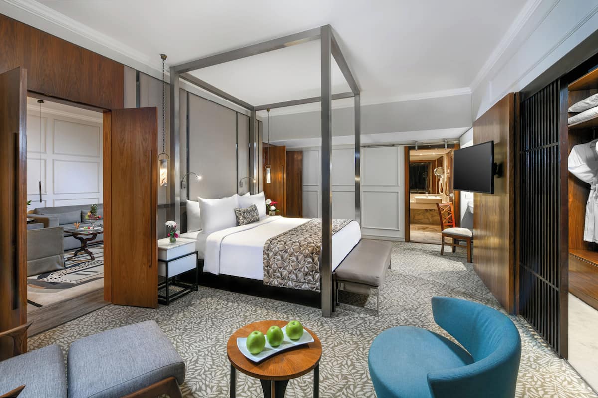 A hotel room with a four-poster bed, a sitting area with a chair and a table, a television, and a bathroom visible through an open door.