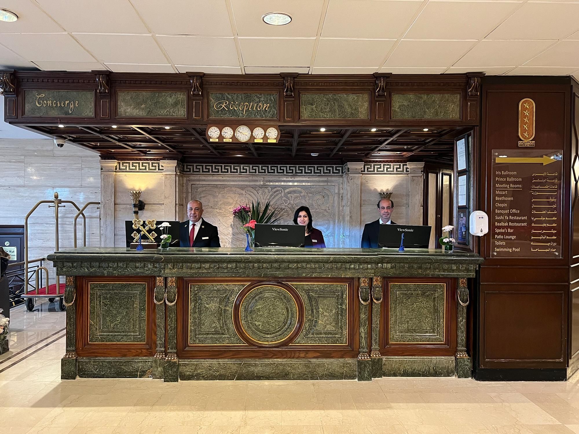 A reception desk with a marble top, two receptionists, and a sign listing hotel amenities.
