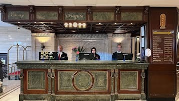 A reception desk with a marble top, two receptionists, and a sign listing hotel amenities.