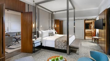 A hotel room with a four-poster bed, a sitting area with a table and chairs, a television, and a bathroom visible through an open door.