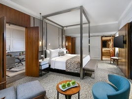 Habitación de hotel con cama con dosel, zona de estar con mesa y sillas, televisor y baño visible a través de una puerta abierta.