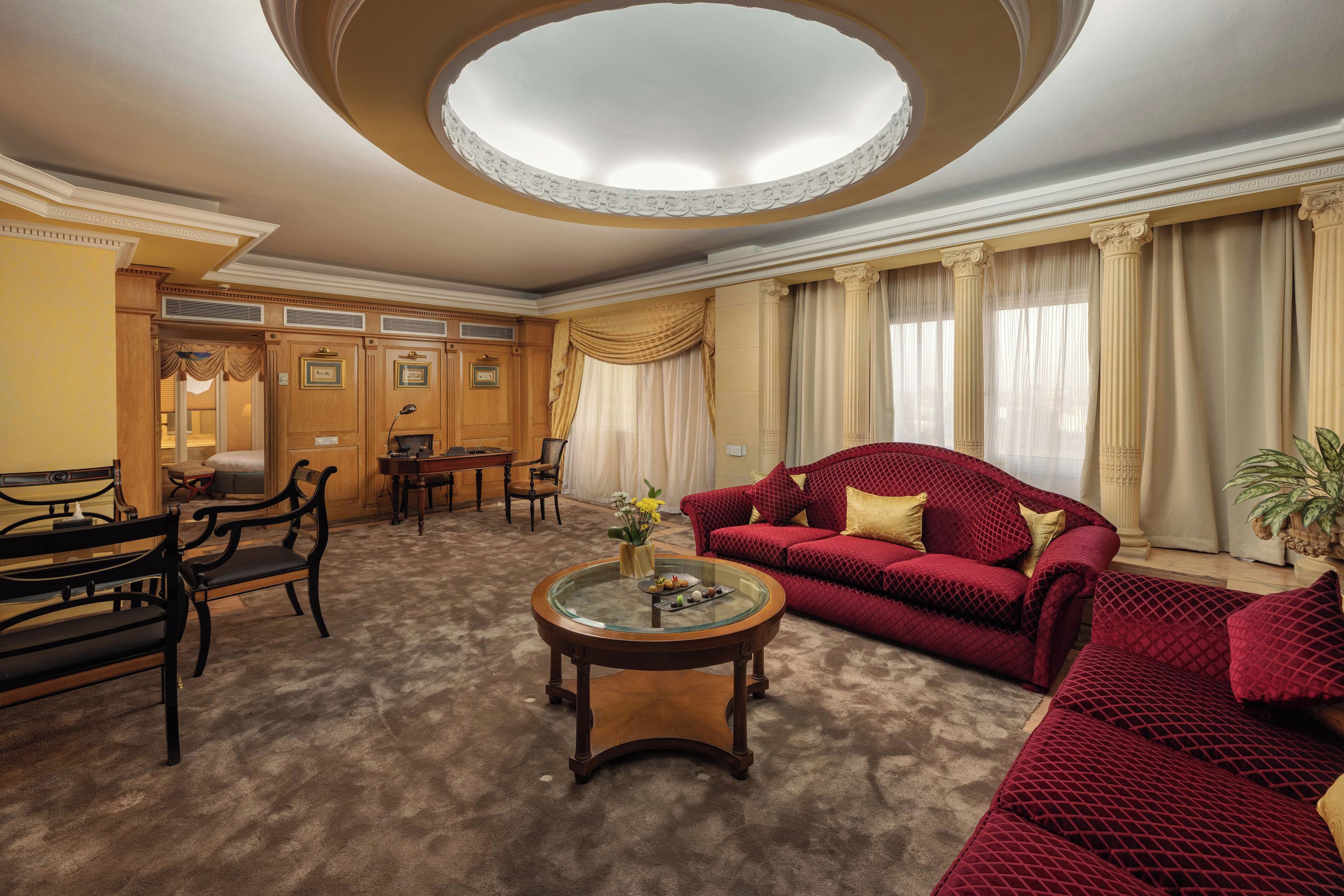 A spacious living room with a red sofa, a round coffee table, and a wooden bench. There is a dining area with a table and chairs, and a large window with curtains.